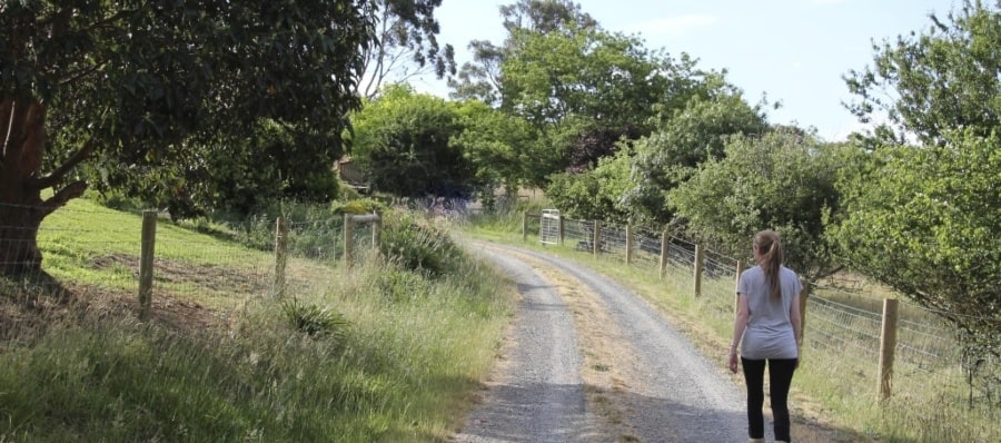 Lady in grey tshirt and black leggings walking away up a long driveway flanked with trees.