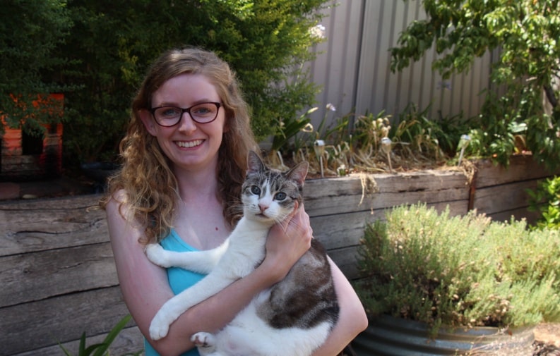 Lady holding cat in foreground, wooden retaining wall with bushes, nectarine tree and lavenders in the background