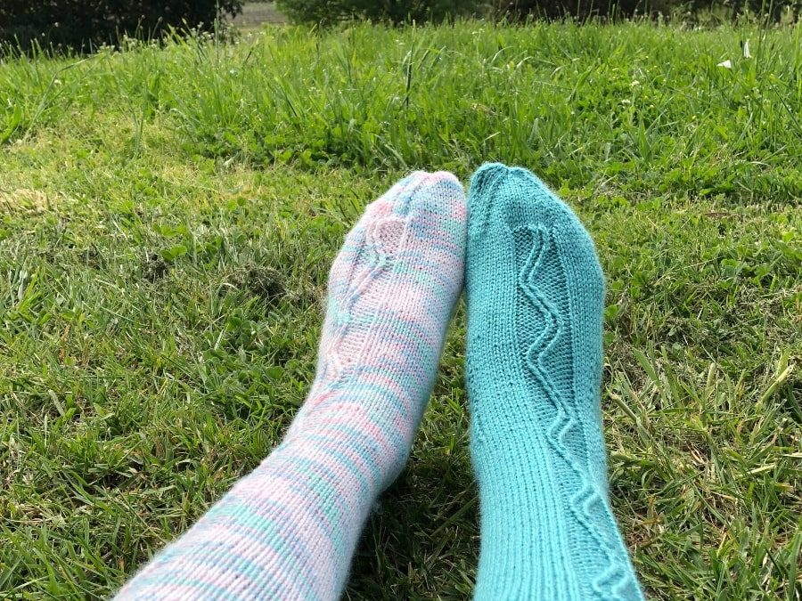 Two legs from knees down with handknitted socks, the left multicoloured and the right in aqua green, sitting on a grass with trees in background.