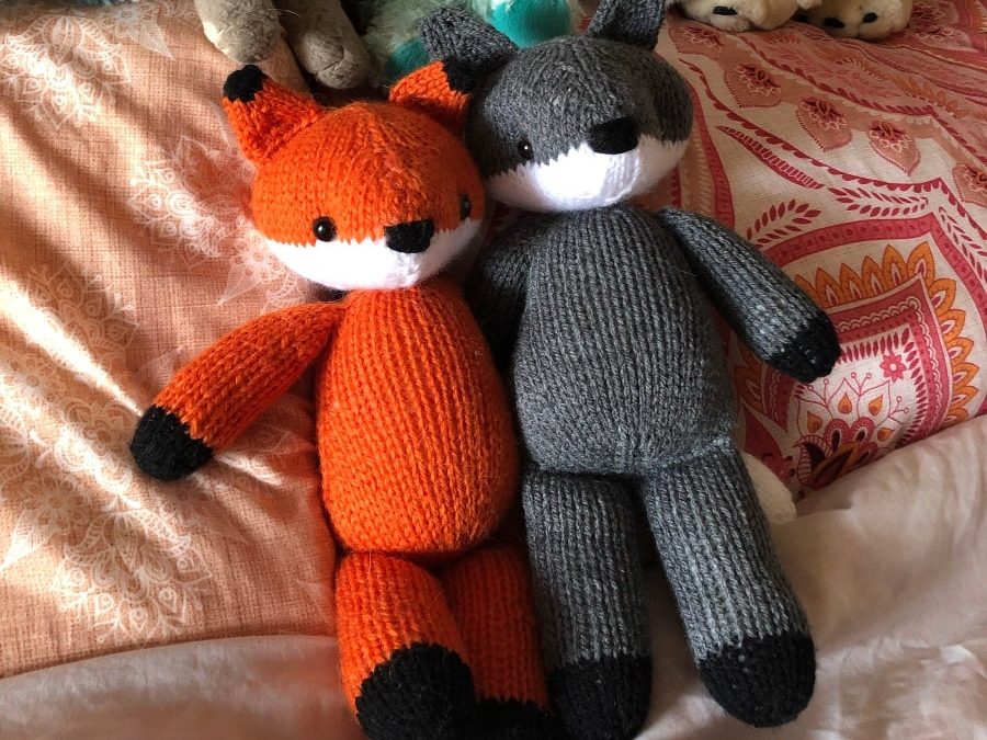 A handknitted fox and wolf sitting together on pink bedspread.