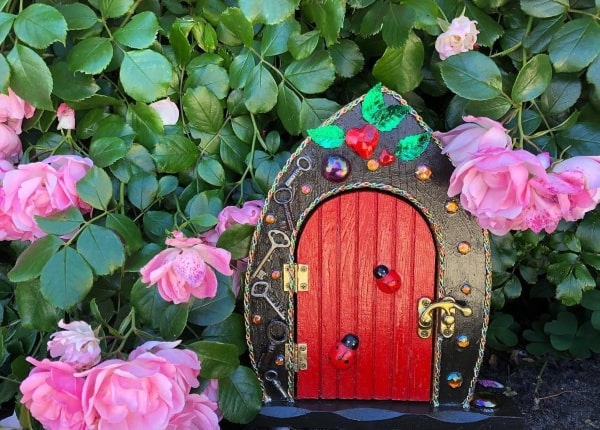 A red decorated fairy door with keys and gems around the frame, and two ladybirds on the red door, set against a background of a pink rose bush.