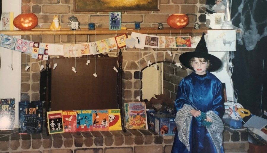 girl dressed as witch in blue cloak beside fireplace with halloween decor