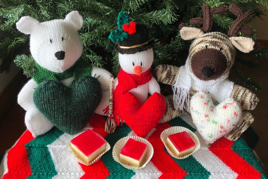 Knitted toys sitting in front of a christmas tree from left to right, a polar bear with a green scarf holding a green heart, a snowman with a red scarf holding a red heart and broom, and a reindeer with a white scarf holding a white heart with red and green speckles on it. Sitting on a knitted rug with green, red and white colour scheme, and three jelly slices.