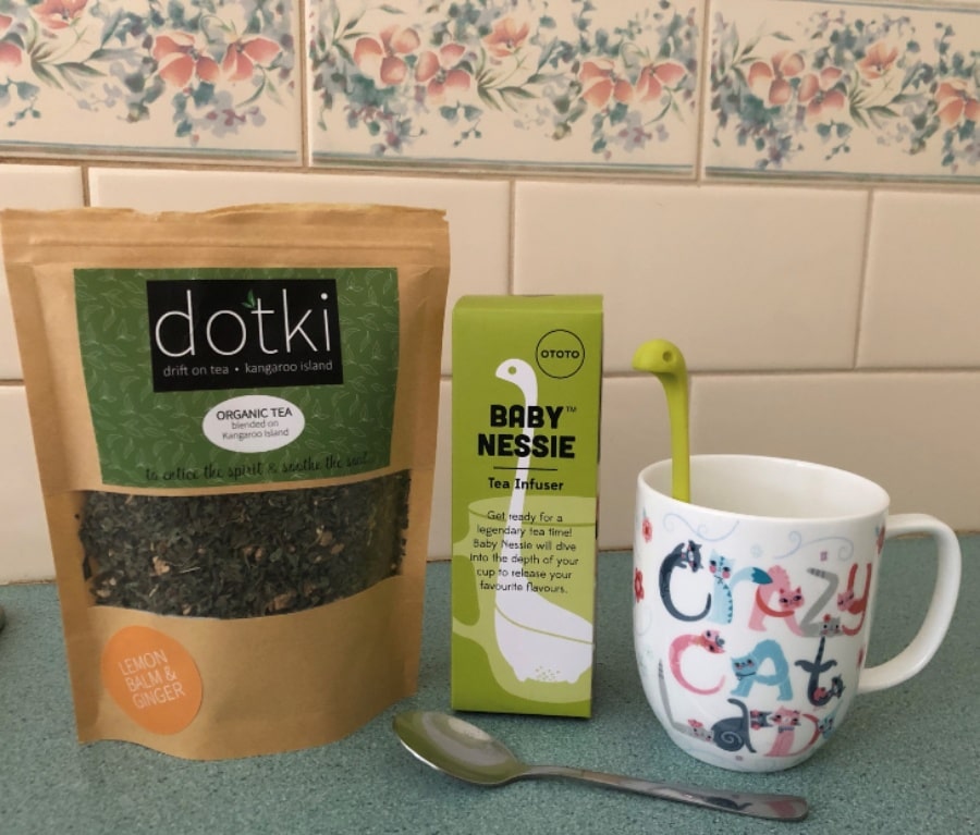 From left to right, a sealable pouch of loose leaf tea from dotki, the box that the baby Nessie tea strainer came in, and baby Nessie in action in a mug that reads "crazy cat lady" set against a cream and floral tiled background.