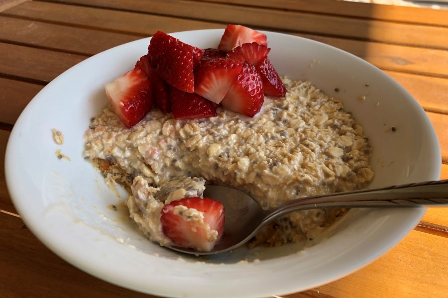 Overnight oat brekkie in a white bowl with strawberries on top and a spoon resting in the oats.